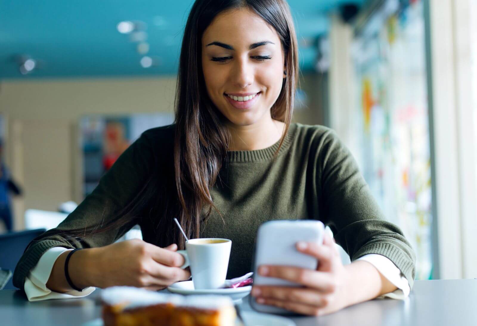 Smiling woman in a cafe enjoying coffee while browsing her phone.