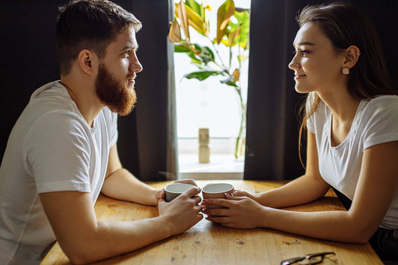 Couple sitting at a table, holding cups and enjoying a conversation together in a cozy indoor setting.