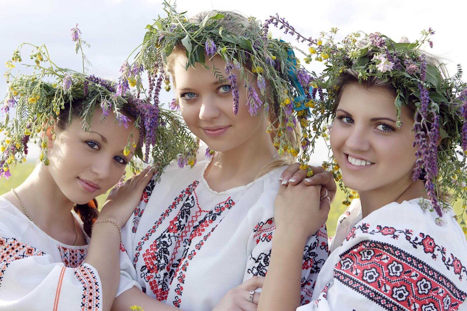 Three smiling young women in Ukrainian embroidered clothing and flower wreaths on their heads.