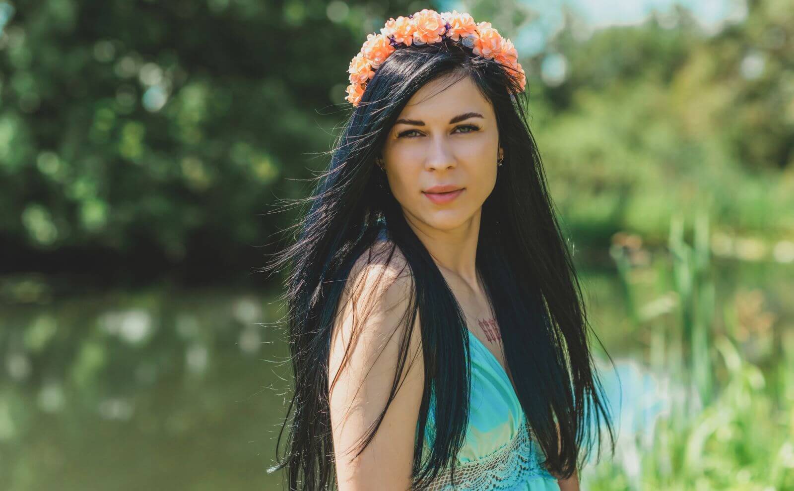Young Ukrainian woman with black hair and floral crown standing near a lake, looking at the camera.